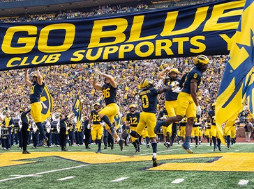 Michigan football players jumping up to hit the GO BLUE sign as they run onto the field.