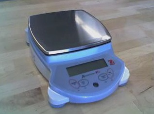 A short, squarish device. The front part has buttons and a simple display panel. The top of the device is a separate metal pan that sits on top of a sensor.