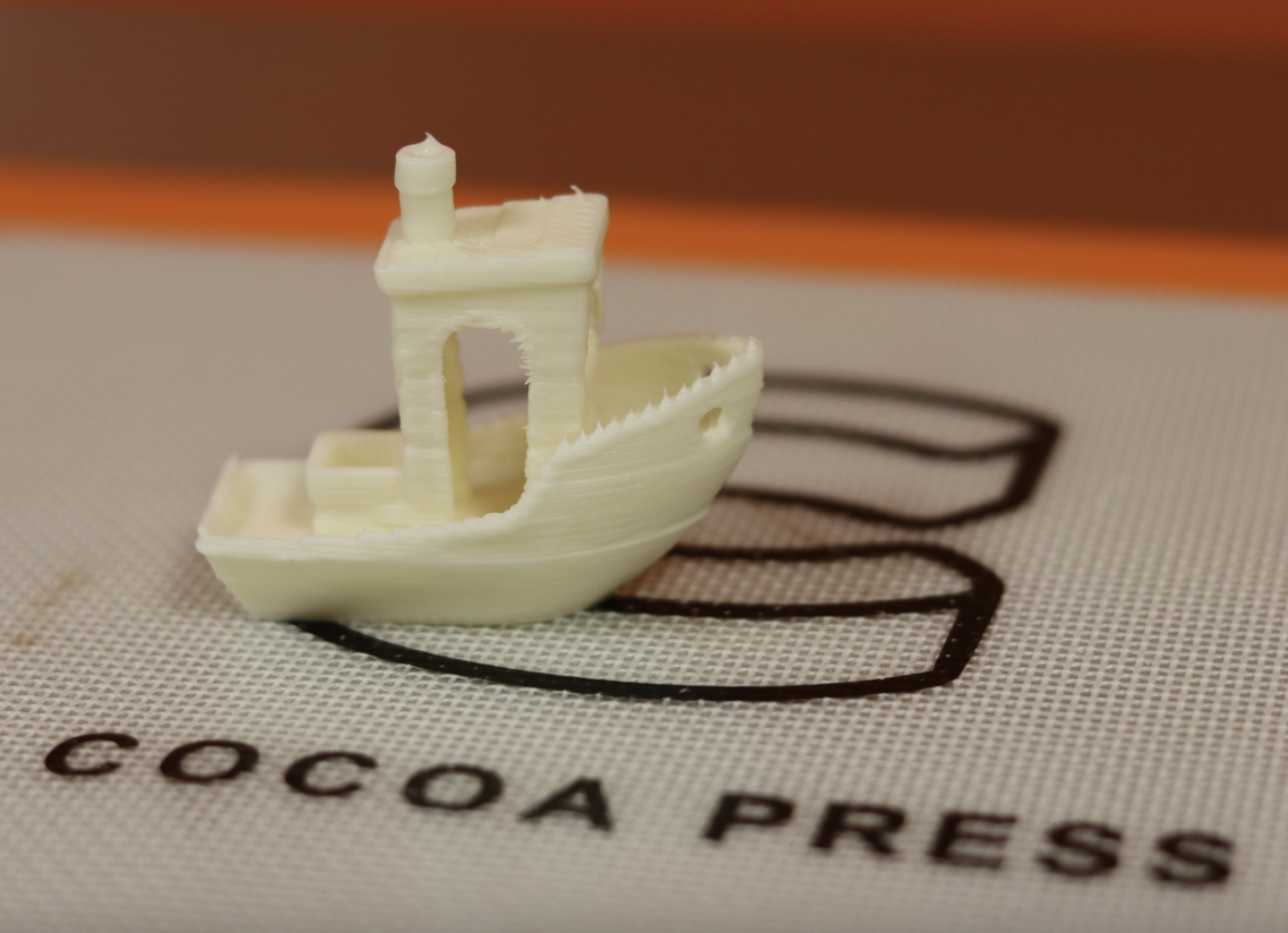 Tiny tug boat made out white chocolate; boat placed on top of a gridded printing surface that has the Cocoa Press logo printed on it in dark brown chocolate.