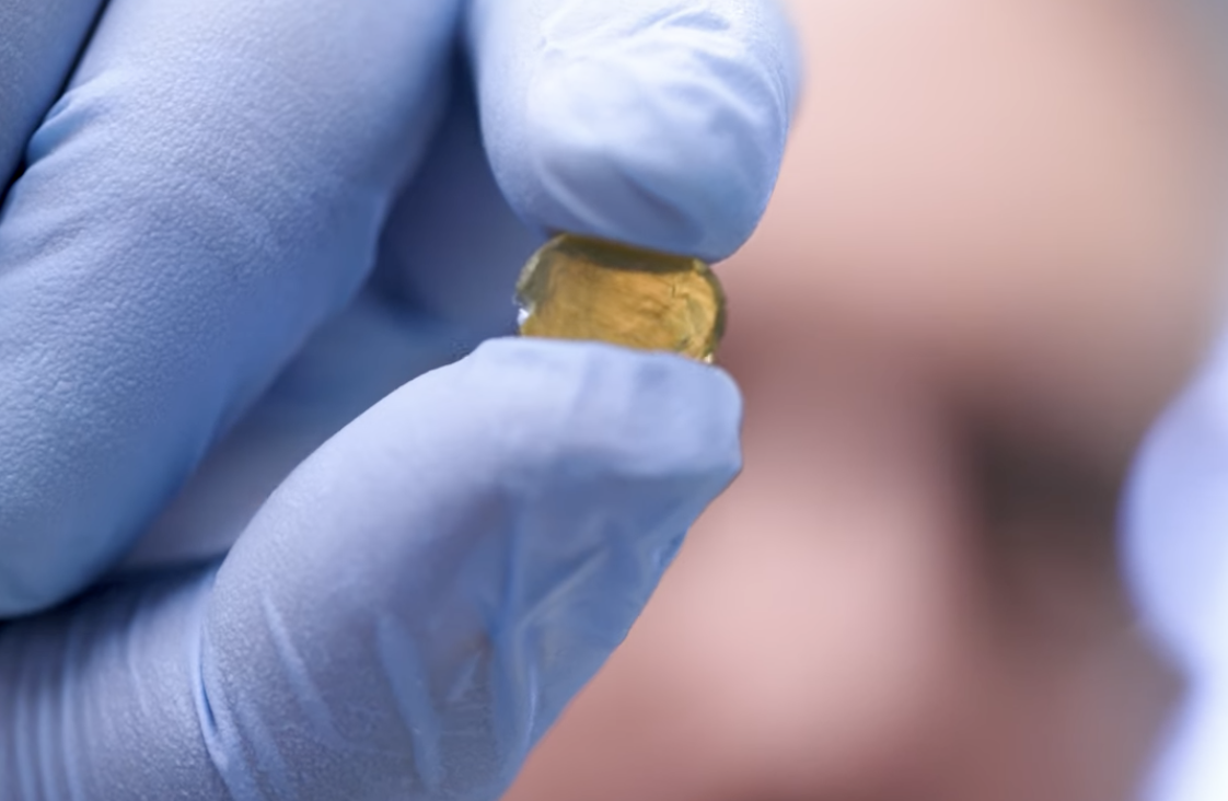 A human hand wearing a blue surgical glove is shown squeezing a transparent yellow disk of gel.