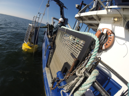 A buoy equipped with solar panels is held over the side of a research ship. The buoy is waiting to be lowered into the water so that it can float around and take measurements.